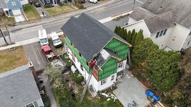 Home Renovation & Remodeling Services in Connecticut. Two‑story house under renovation with green exterior sheathing, workers on the roof, ladders, and construction vehicles in the driveway.