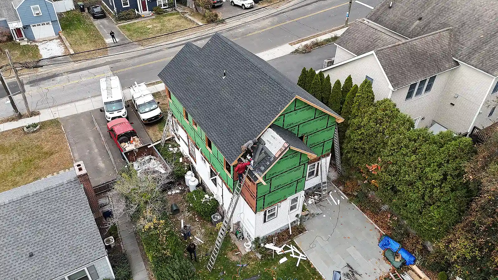 Home Renovation & Remodeling Services in Connecticut. Two‑story house under renovation with green exterior sheathing, workers on the roof, ladders, and construction vehicles in the driveway.