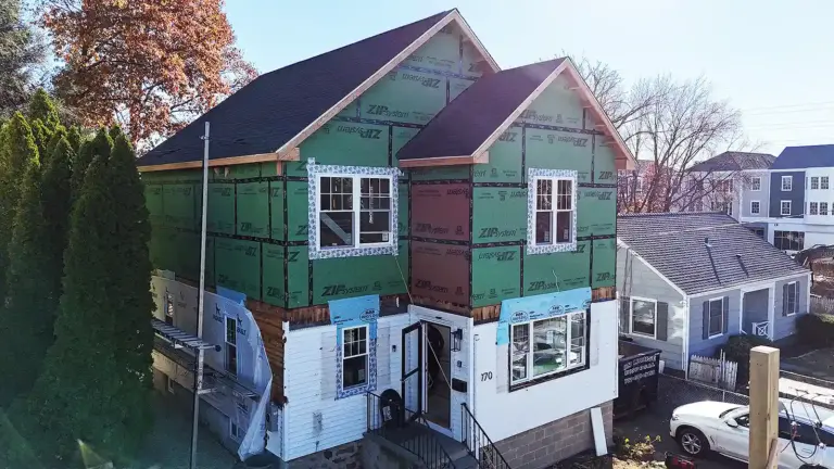 Home renovation and remodeling in Connecticut. House under renovation with new upper-level framing covered in green and red sheathing, windows installed, and construction materials around the yard.